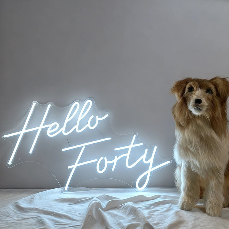 A glowing white neon-style sign that reads 'Hello Forty' sits on a white bed against a gray wall, with a fluffy brown-and-white dog standing to the right.