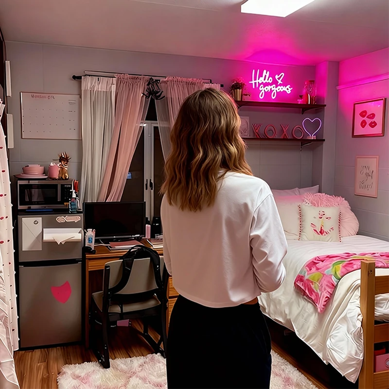 Person standing in a bedroom with pink neon sign, desk, and bed.