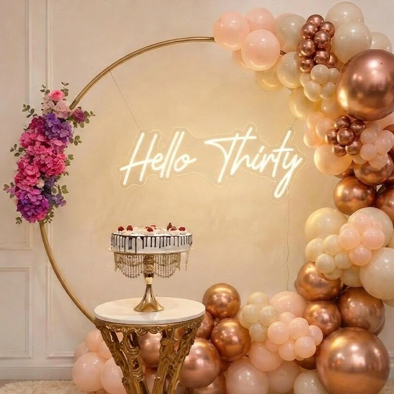 A decorative party backdrop with a large gold arch, blush pink, cream, and metallic rose-gold balloons, a neon-style 'Hello Thirty' sign, and a small cake on a pedestal stand.