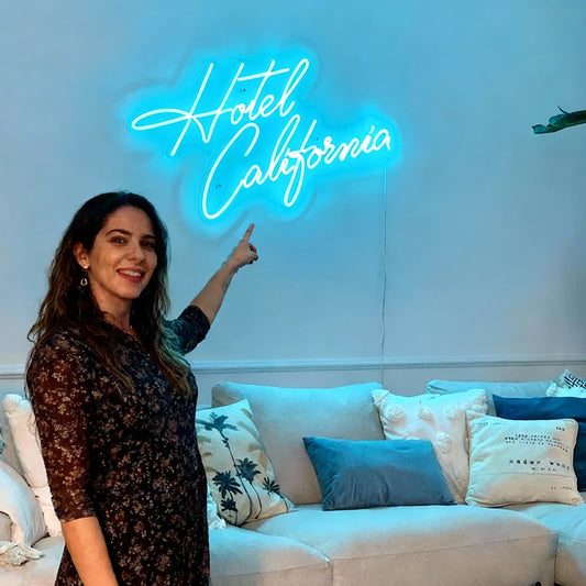 Woman pointing at a neon sign that reads 'Hotel California' in a room with a couch and pillows.