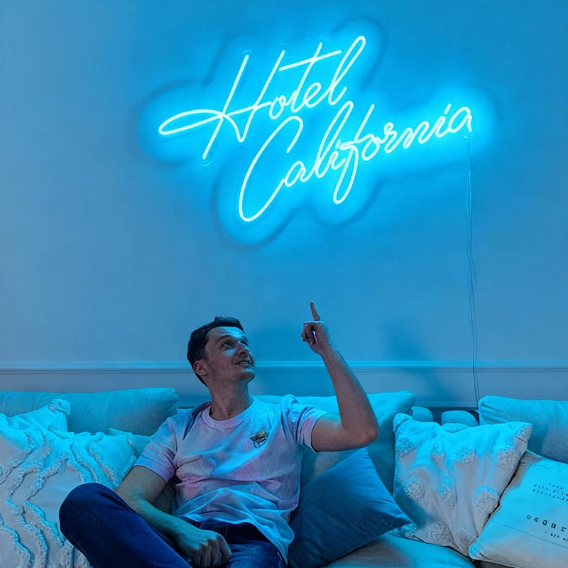 Man sitting on a couch pointing at a neon 'Hotel California' sign.