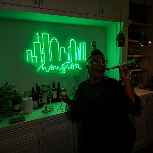 Person celebrating indoors with a champagne glass and party blower in front of a green neon "Houston" skyline sign, with a countertop featuring wine bottles and decorative plants.