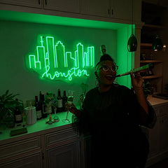 Person celebrating indoors with a champagne glass and party blower in front of a green neon "Houston" skyline sign, with a countertop featuring wine bottles and decorative plants.