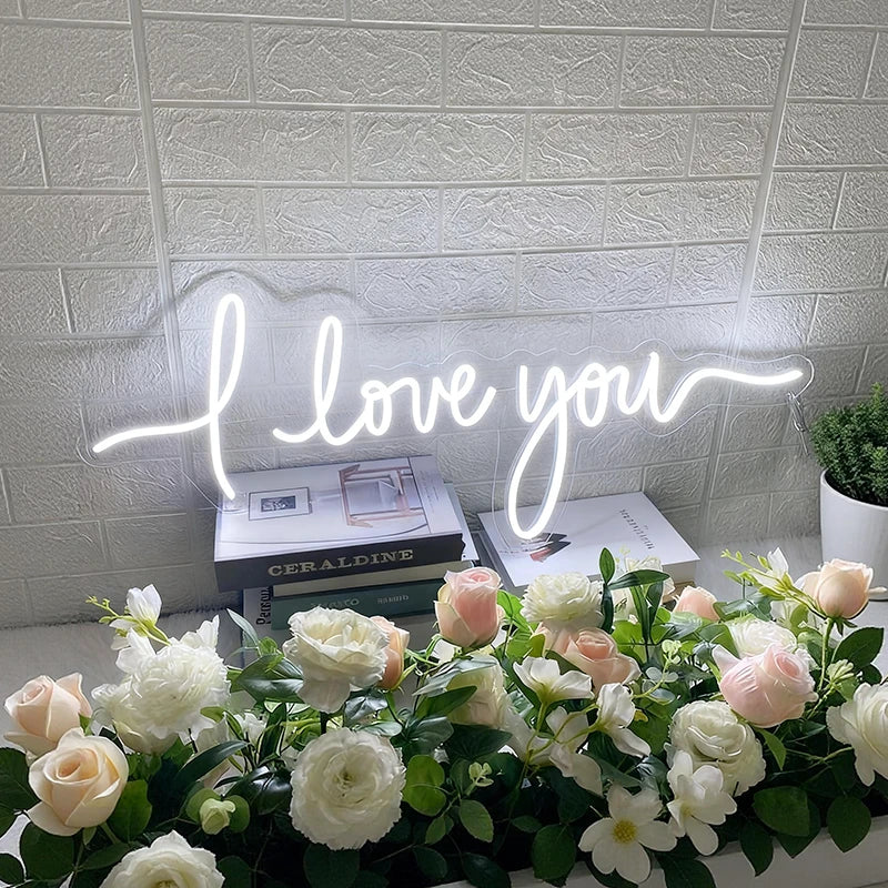 Neon sign saying 'I love you' above a table with flowers and books.