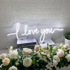 Neon sign saying 'I love you' above a table with flowers and books.