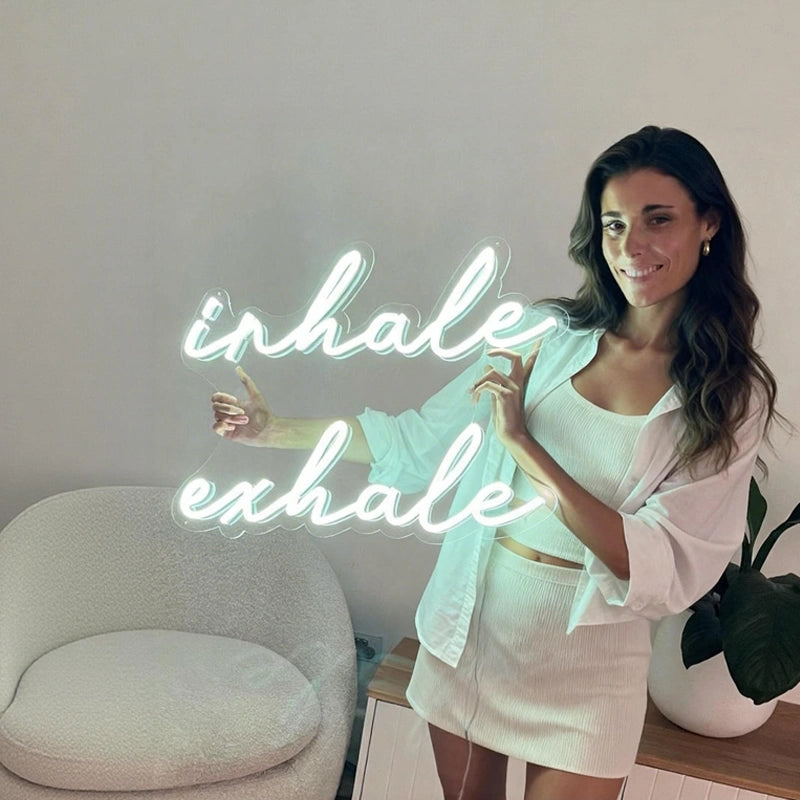 Woman in a white outfit standing next to a neon sign that reads 'inhale exhale' in a minimalistic room.