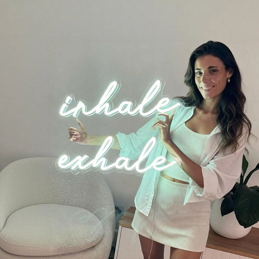 Woman in a white outfit standing next to a neon sign that reads 'inhale exhale' in a minimalistic room.