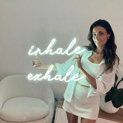 Woman in a white outfit standing next to a neon sign that reads 'inhale exhale' in a minimalistic room.