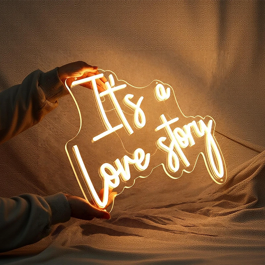 A person holds a glowing acrylic sign that reads “It’s a Love story” in warm white light against a softly lit fabric backdrop.
