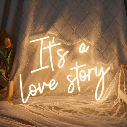 A glowing warm white neon sign that reads 'It's a love story' placed on draped fabric with a warm light radiating across the scene.