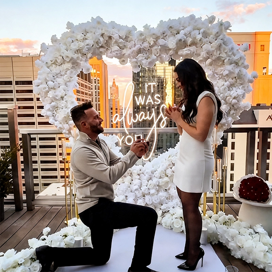 Man proposing to a woman in front of a floral arch with 'It was always you' neon sign on a rooftop.