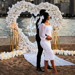 Couple in white attire standing under a floral arch with 'it was always you' neon sign on a beach.