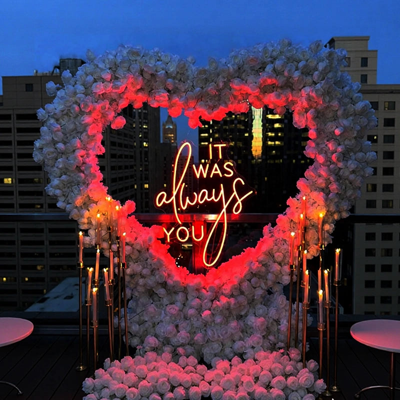 Neon sign in a heart shape surrounded by flowers with 'It was always you' text, set against a city skyline at night.