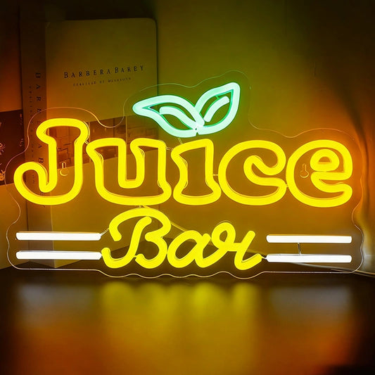 A glowing LED neon sign that reads "Juice Bar" in a stylized yellow font. Above the word "Juice" is a small green icon of two leaves. The word "Bar" is centered between two sets of horizontal white lines. The sign is mounted on a clear acrylic backing and is shown illuminated against a dark background next to a book.