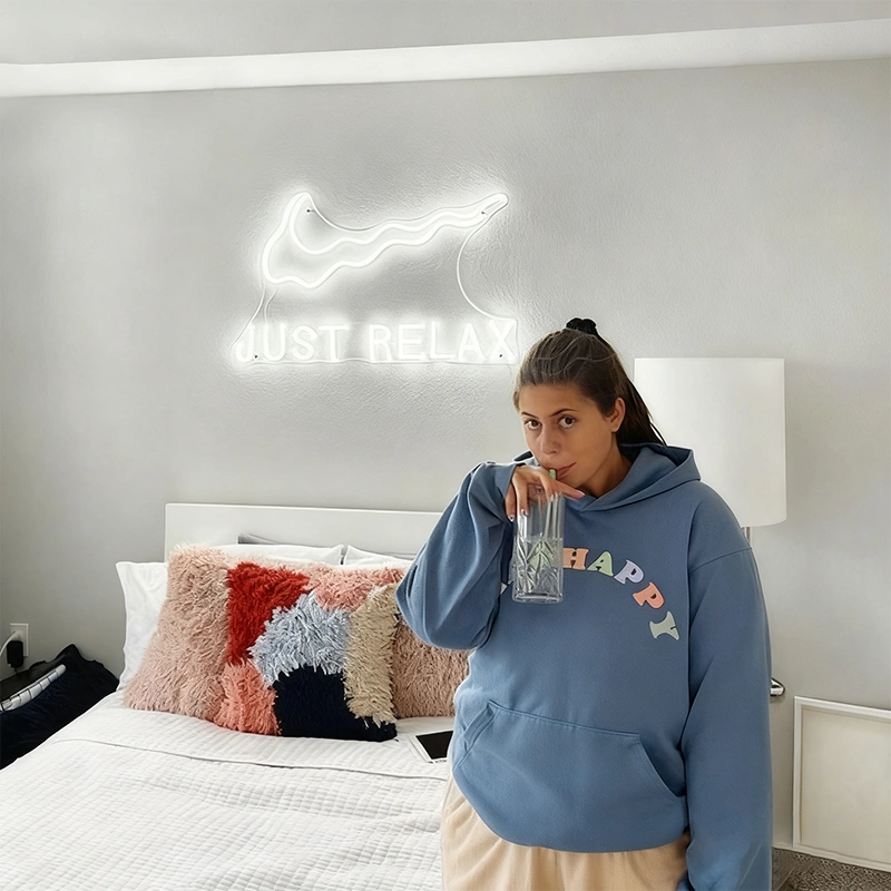 Person wearing a blue hoodie with colorful text in a bedroom with a 'just relax' neon sign.