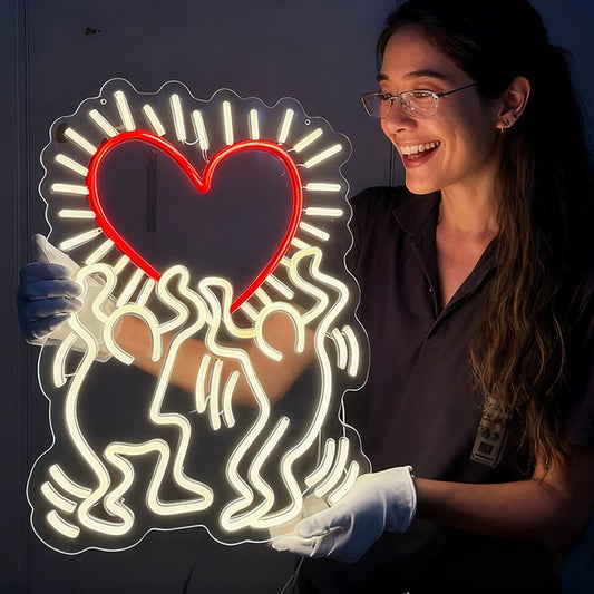 A woman wearing glasses, white gloves, and a dark uniform smiles while holding a large illuminated wall sign. The sign features a red heart outline surrounded by white abstract line art keith haring love on a dark background.