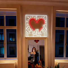 A square illuminated wall sign featuring a glowing red heart and white outline figures inspired by Keith Haring, mounted above a doorway in a warmly lit interior. A person is visible taking a mirror selfie in the reflected room below.