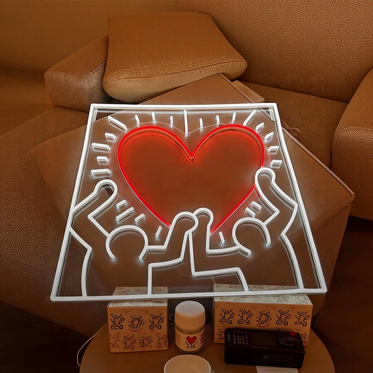 A square LED neon sign featuring a glowing red heart and white outline figures inspired by Keith Haring, displayed on a brown leather sofa with tissue boxes and a small jar underneath.