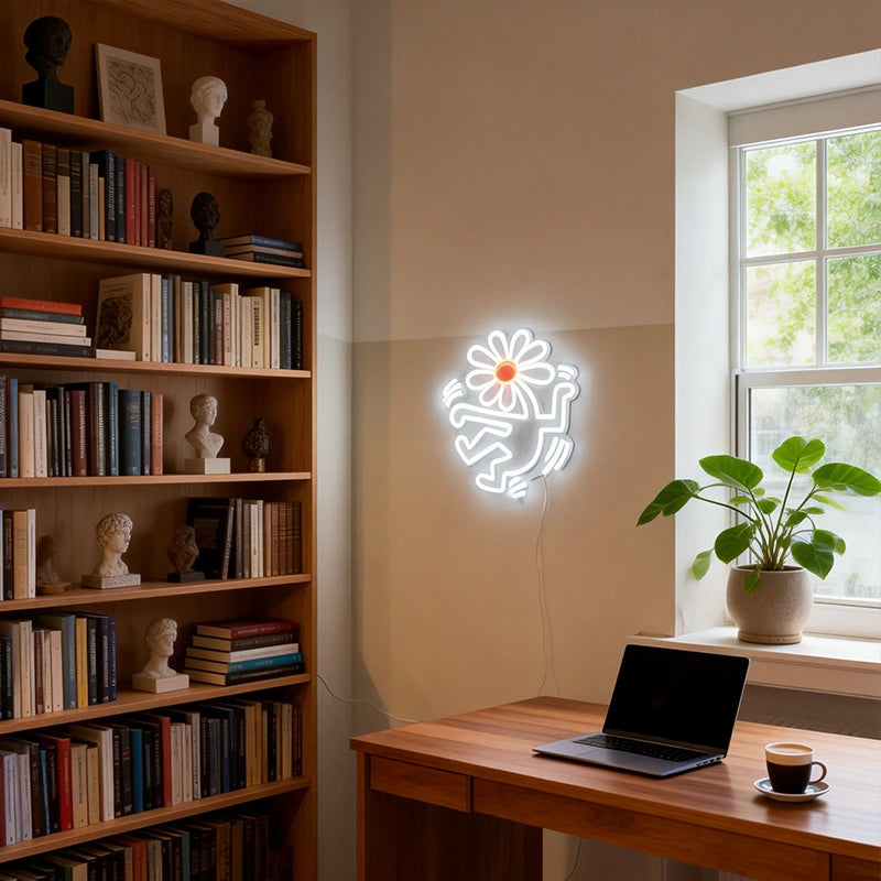 Neon light shaped like a flower head with human body on a wall next to a bookshelf and desk with a laptop and coffee.