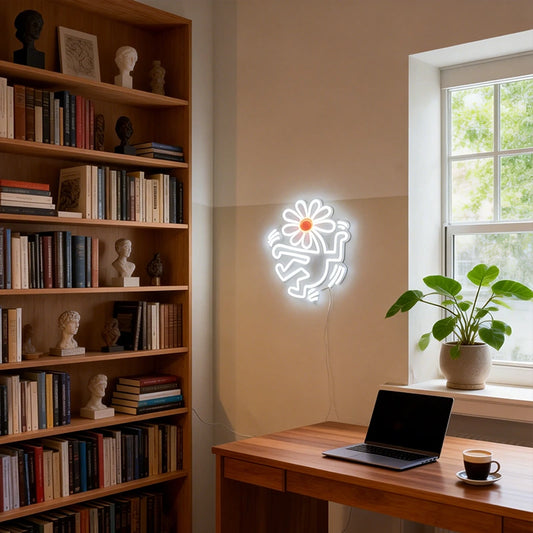 Neon light shaped like a flower head with human body on a wall next to a bookshelf and desk with a laptop and coffee.