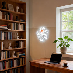 Neon light shaped like a flower head with human body on a wall next to a bookshelf and desk with a laptop and coffee.