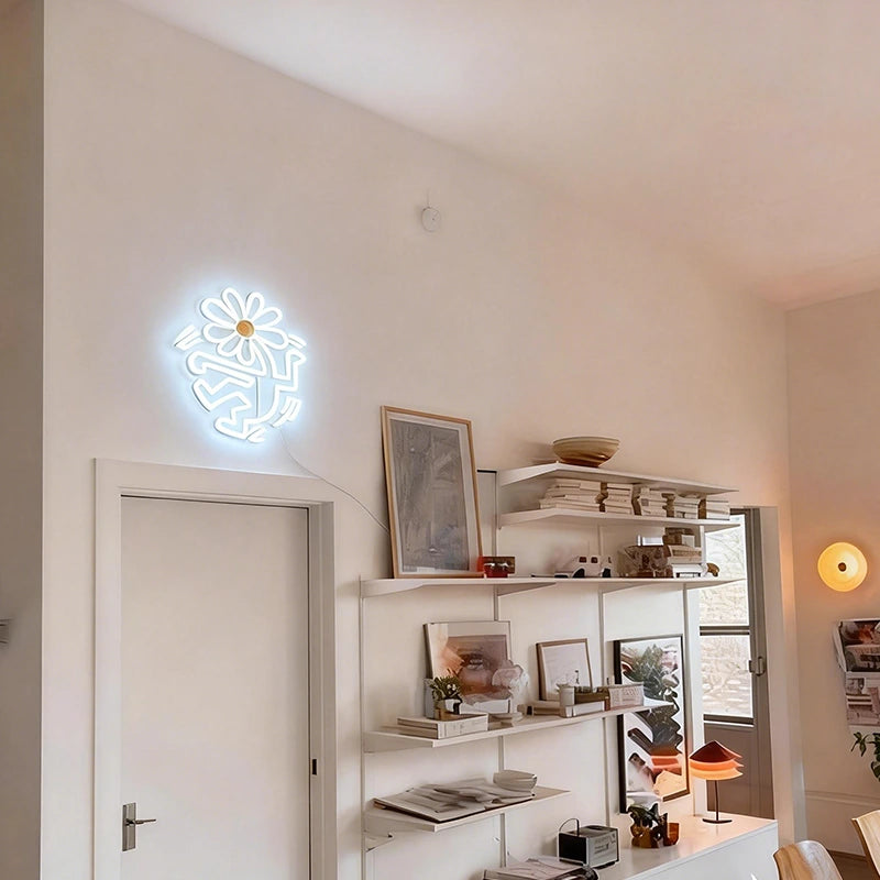 Neon flower head light on a wall above a shelf with decor items in a room.