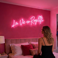 Woman sitting on a bed in a room with a light pink neon sign reading 'La Vie en Rose' on a wall.