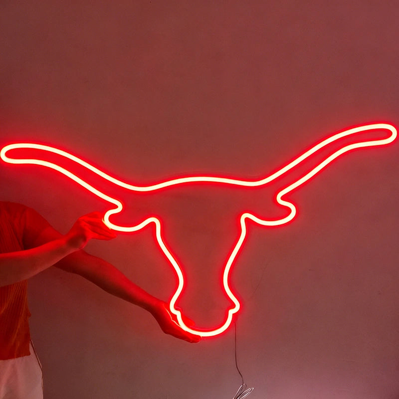 A red neon light sign shaped like a longhorn steer or Texas longhorn outline, shown against a dark wall with a person's hands and arms holding it in place.