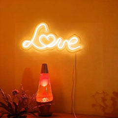 Neon sign spelling 'LOVE' on a warm white wall with a lava lamp and plant below.