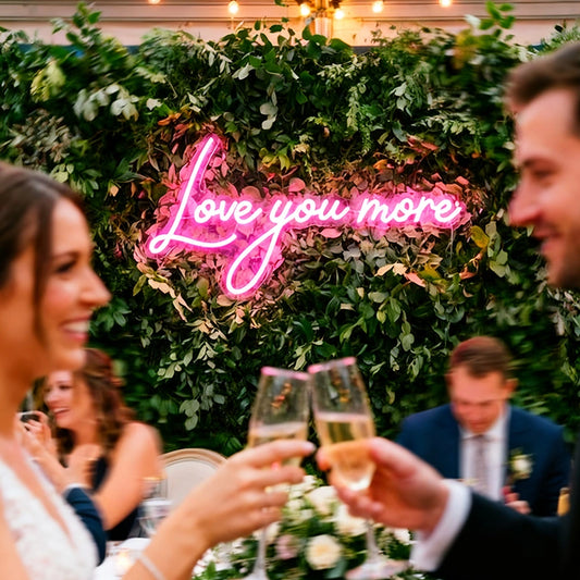 A wedding reception scene with a green leaf wall backdrop featuring a hot pink neon sign that reads 'Love you more.' A bride and groom are in the foreground clinking champagne flutes, with guests and warm string lights visible in the background.