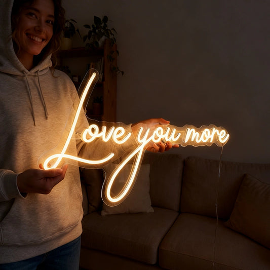 A woman in a light gray hoodie holds a glowing LED neon sign that reads 'Love you more' in a warm white light, set against a dimly lit living room background with a sofa.