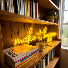 A glowing lemon yellow neon sign that reads 'make it happen' mounted on a wooden bookshelf next to a window, with books, a mug, and glasses around it.
