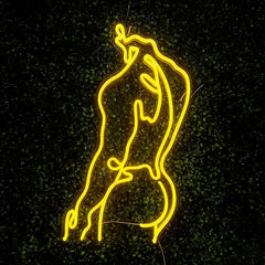 Lemon yellow neon line art sign of a stylized human figure against a dark leafy background, glowing brightly in the night.