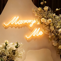 Neon sign with 'Marry Me' surrounded by flowers on a light background