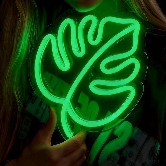 A person holds a glowing green neon sign shaped like a monstera leaf against a dark background.