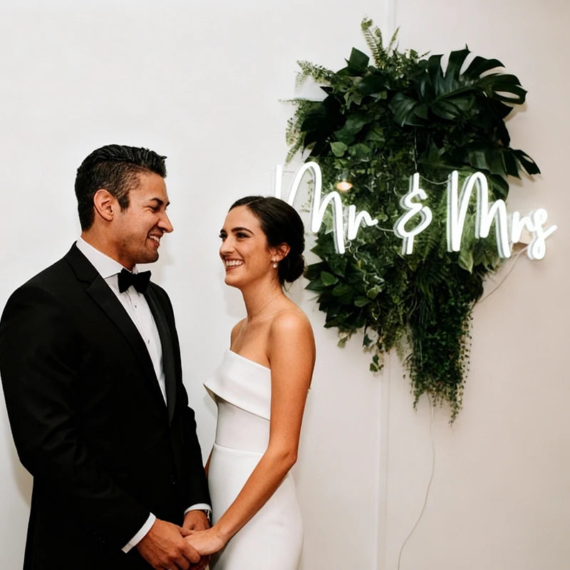 Man and woman in formal attire standing in front of a 'Mr & Mrs' sign with greenery.