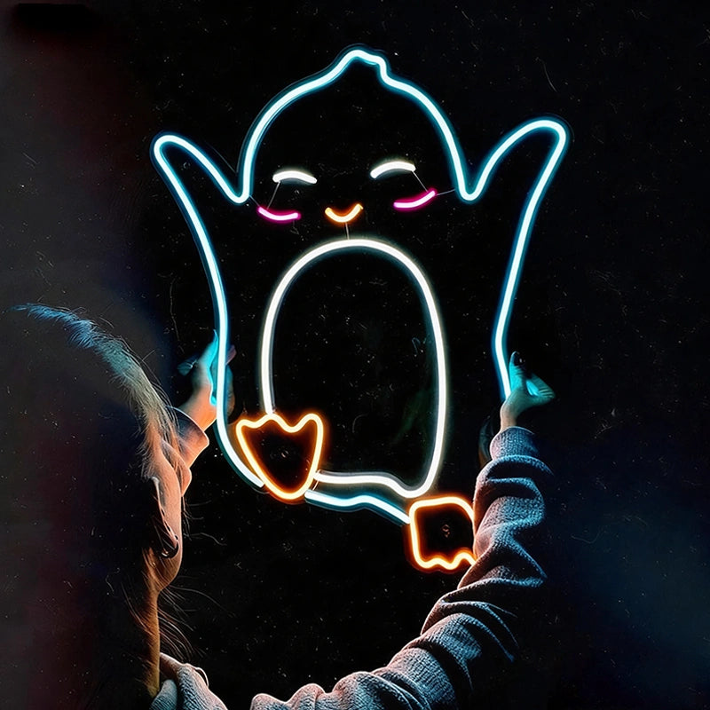 A person in a gray hoodie holds up glowing neon light shape like a penguin against a dark background. The neon design glows in ice blue, white, hot pink, and orange.
