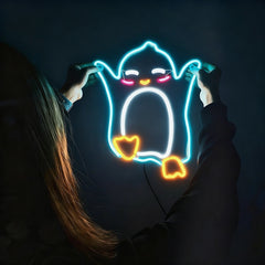 A person holds up a glowing neon sign shaped like a cartoon ghost penguin, with bright blue, white, hot pink, and orange light against a dark background.