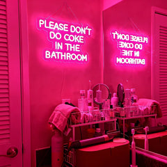 Neon sign warning 'Please don't do coke in the bathroom' in a pink-lit room with bathroom items.