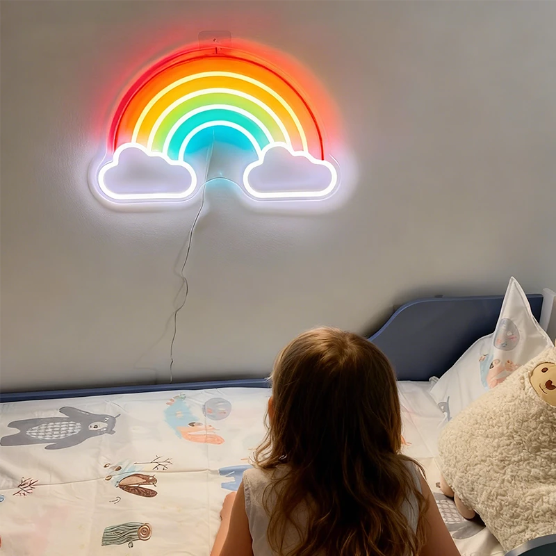 Child sitting on a bed looking at a rainbow-shaped neon light on the wall.