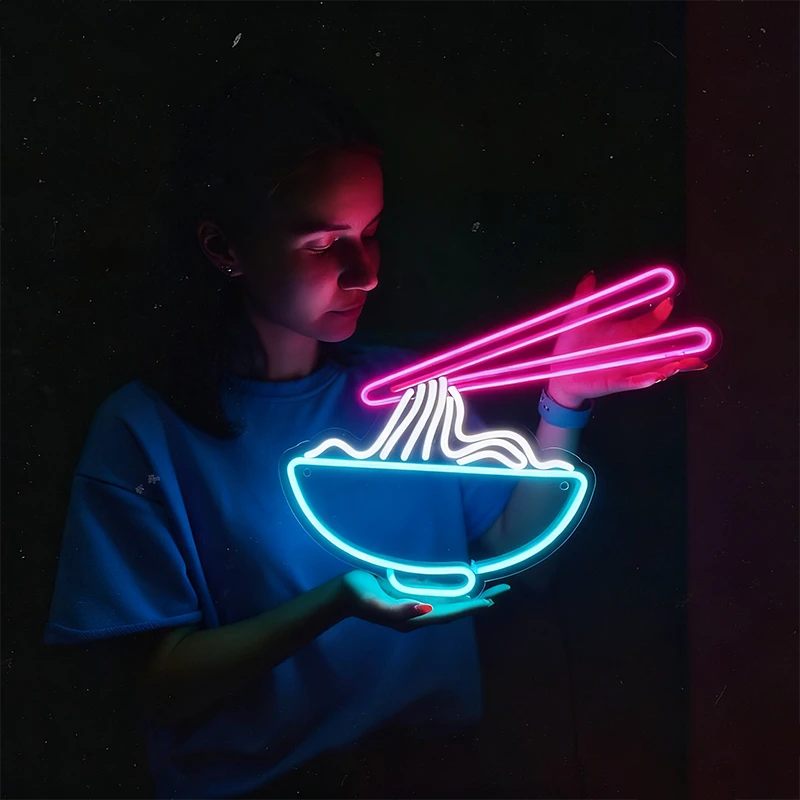 Person holding a neon sign of a bowl of noodles with chopsticks against a dark background