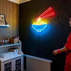 Neon sign of a bowl of noodles with chopsticks on a dark wall, person in red shirt standing to the right.