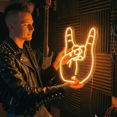 A man in a black studded leather jacket holds a glowing orange neon sign shaped like a hand with a rose, set against a dark acoustic foam wall with guitars hanging in the background.