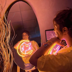 Person holding a sailor cat neon sign looking at herself in a mirror with a pinkish hue.