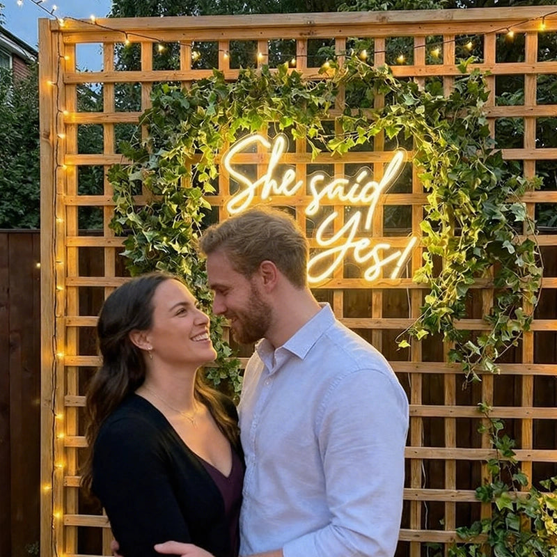 A smiling couple standing in front of a wooden lattice backdrop decorated with green ivy and warm string lights, with a glowing neon sign reading “She said Yes!” behind them.