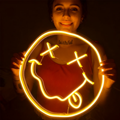 Person holding a neon sign shaped like a smiley face with a cross inside, glowing warmly.