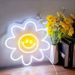 Neon flower-shaped light with a smiley face next to a vase of lemon yellow flowers on a wooden surface.