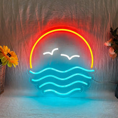 A neon-style wall light shaped like a sunset over water, with orange sun arc, white seagulls, and ice blue waves, displayed against a wrinkled fabric backdrop with decorative flowers at the sides.