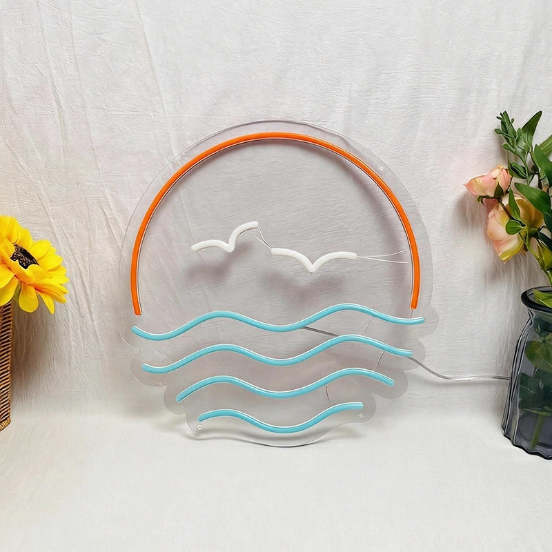 A decorative acrylic LED light shaped like a beach sunset, featuring a clear circular frame with an orange arch, two white seagull silhouettes, and ice blue wavy lines representing the ocean. The light is shown on a white fabric backdrop with decorative yellow flowers on either side.