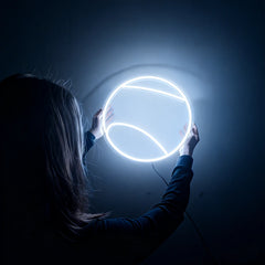 Person holding a round white tennis ball neon light against a dark background